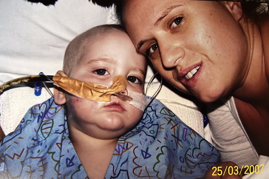 A young boy with no hair and a tube in his nose sits in a hospital bed with his mum beside him
