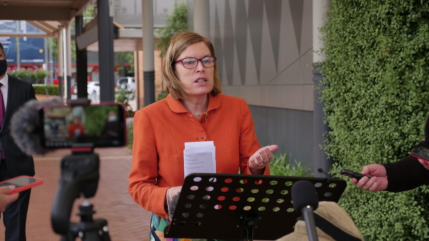 woman with glasses and orange jacket addresses media