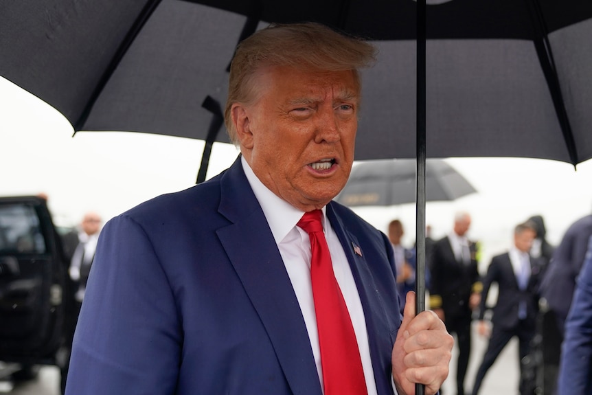 Donald Trump, wearing a red tie and blue suit, holds up a dark black umbrella
