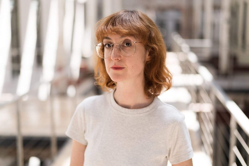 Wendy Syfret is standing in a very bright large room and looking at the camera. She has glasses on and a pale t-shirt.