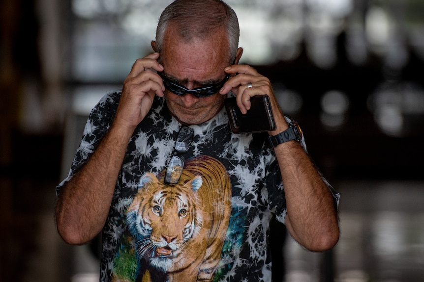 A white man wearing sunglasses and a printed T-shirt looks down as he leaves court