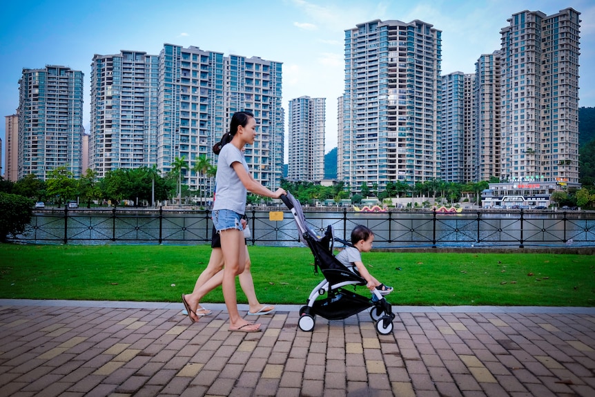 A woman pushes a baby in a pram past a row of high rise residential buildings 