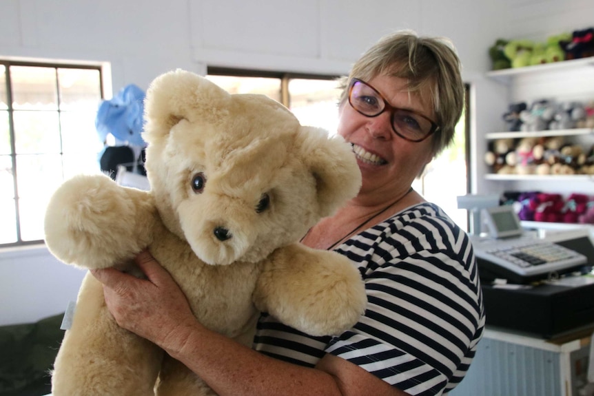 A middle-aged woman with short blonde hair and glasses holds up a large honey-coloured teddy bear.