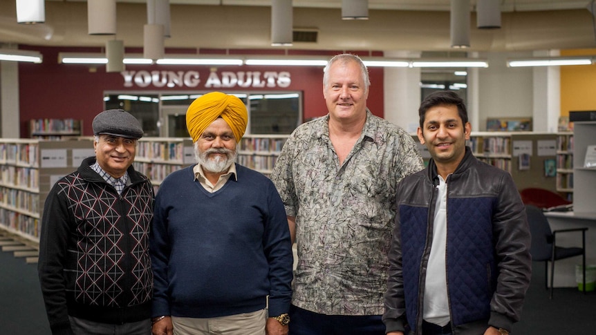 Image of four men standing in a row in a public library, with shelves of books in the background.