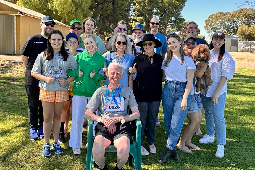 an elderly man in a green chair sitting in front of a large group of men and women, one holding a dog