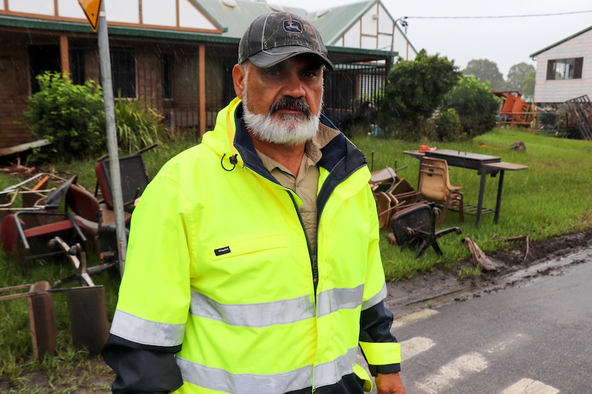 A middle-aged man with a white beard stands in front of a flood-affected house. he is wearing high-vis gear.