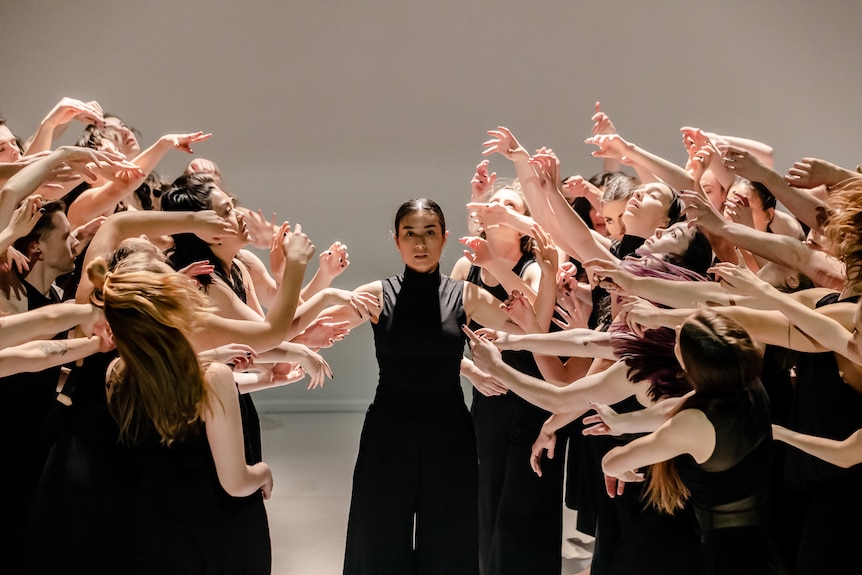 On stage a single dancer in black stands in the middle of a crowd of dancers divided into two, their arms and hands reach out