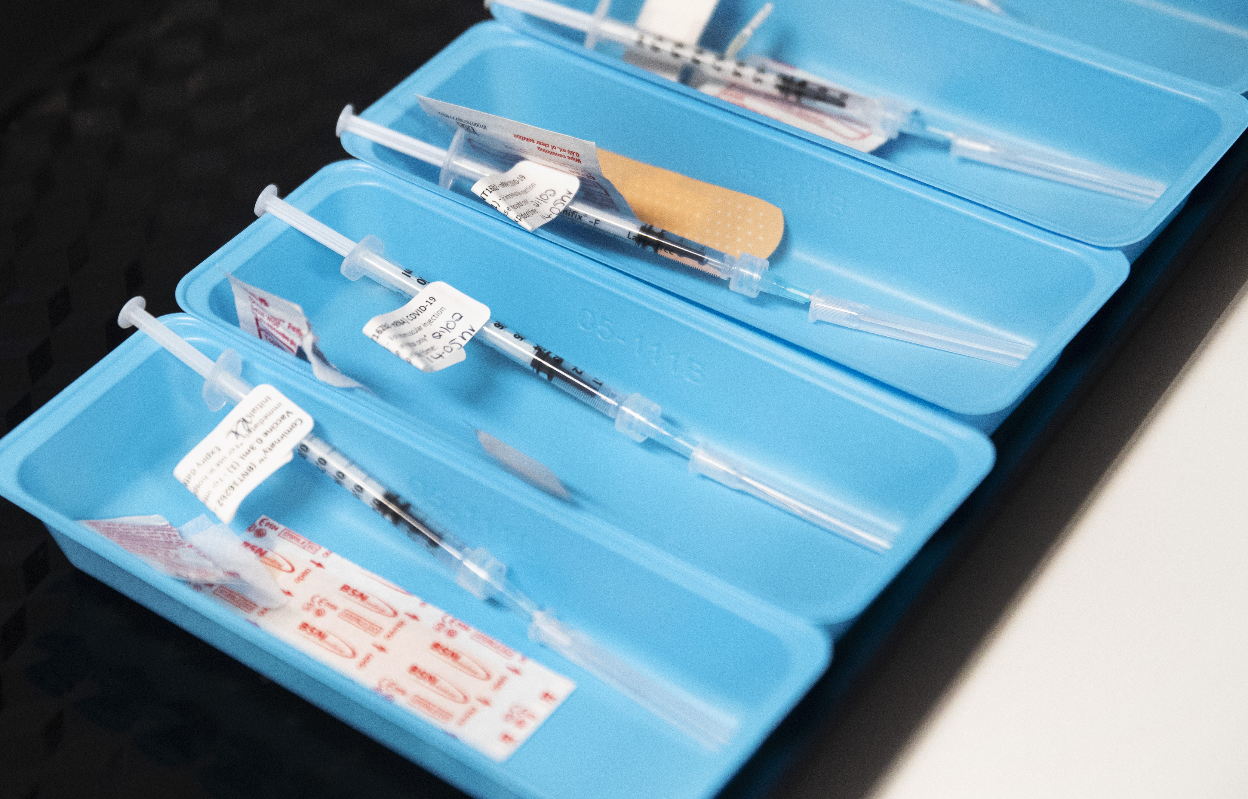 Small blue trays lined up, each with a single syringe of COVID-19 vaccine and a bandaid at a vaccination centre.