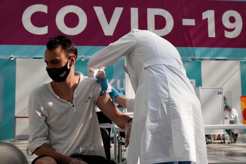 흰색 실험실 가운과 장갑을 입은 의료 종사자가 마스크를 쓰고 앉아 있는 남자에게 백신을 줍니다.