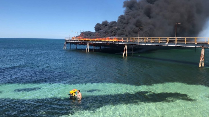Two men in the sea with a jetty in flames behind