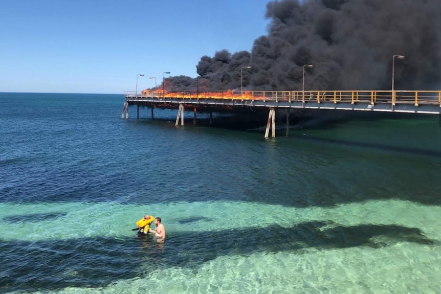 Two men in the sea with a jetty in flames behind