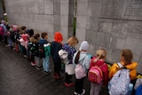 Children wearing backpacks bow their heads down at a war monument in Warsaw, Poland.