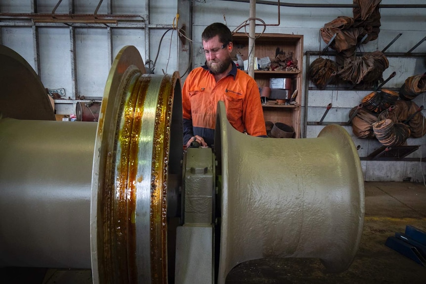 A man in orange hi-vis shirt and protective eye and ear equipment repairs a large metal component of a ship