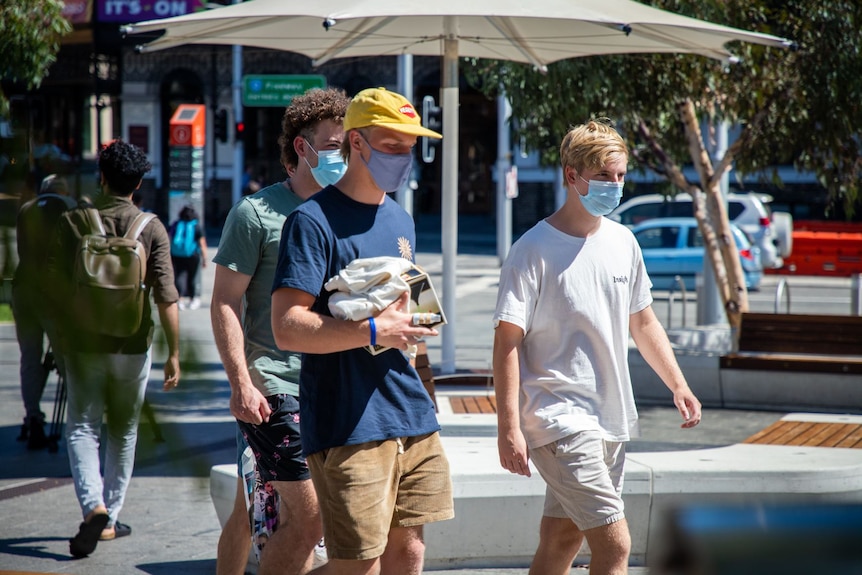 Young men walk in city sunshine wearing masks.
