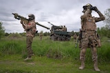 A truck sits in a field as a soldier aims a shoulder-mounted rocket laucher, while another looks through binoculars.