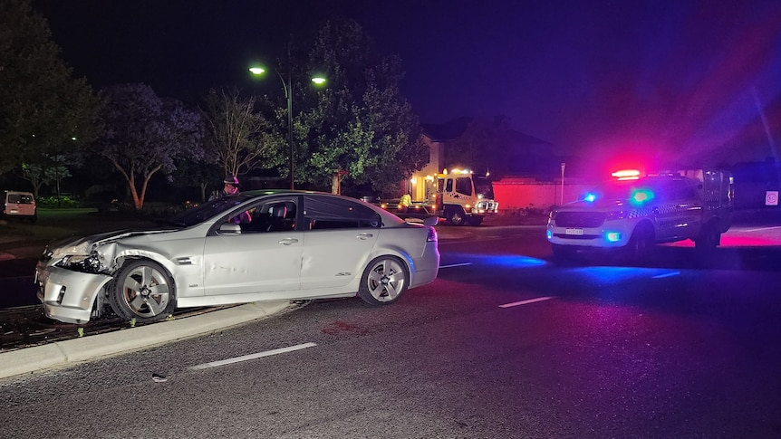 A damaged silver car, with a police car parked behind it.