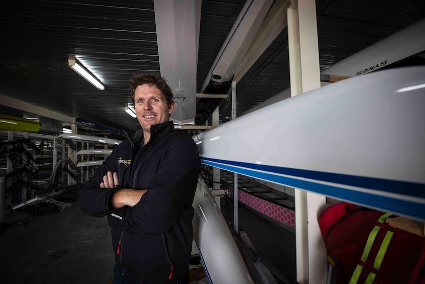 A man folds his arms and smiles as he poses for a photo in front of a boat in a rowing shed
