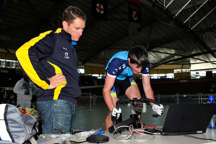 A man watches as a young cyclist rides on a stationary bike.