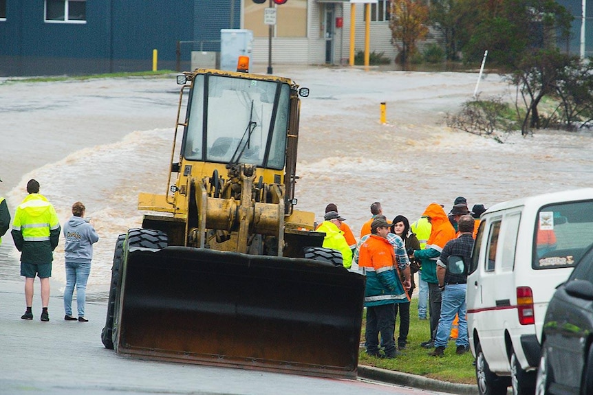 A bulldozer sits next to a flooded road as emergency services workers stand by.