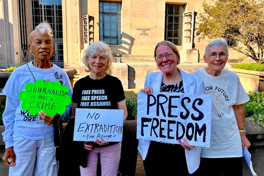 Four women hold signs with messages like 'press freedom' and 'no extradition' in front of a brick building.