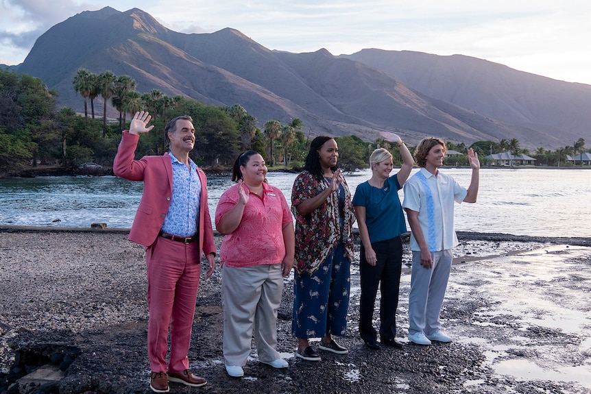 Five people stand waving on a dark beach, with the ocean, palm trees and mountains in the background.