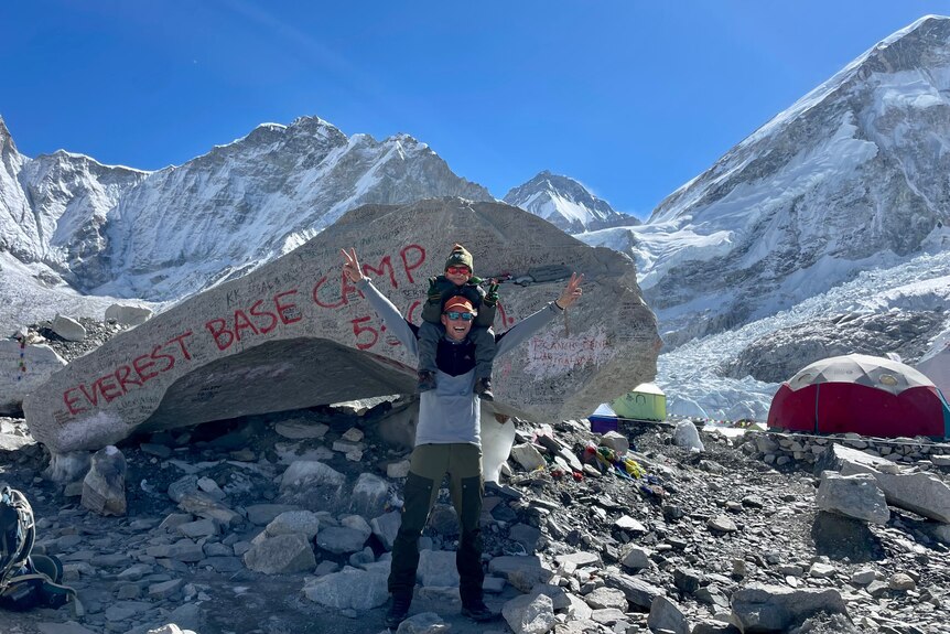 A boy sits on his dads shoulders, with arms in the air in front of a boulder that says Everest Base Camp. 