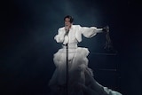 A young man wearing a big white dress stands holding a microphone and singing at Eurovision.