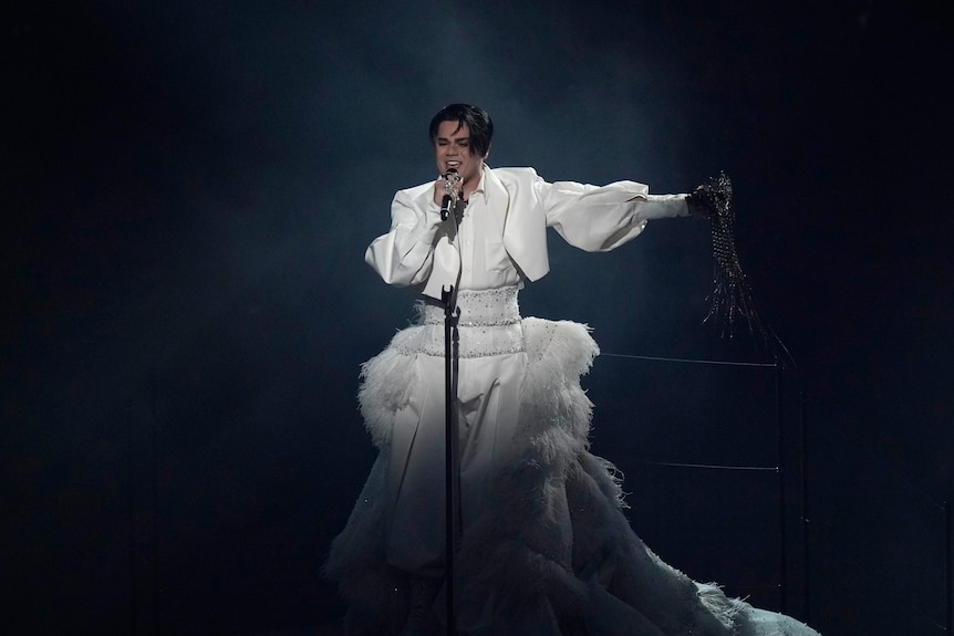A young man wearing a big white dress stands holding a microphone and singing at Eurovision.