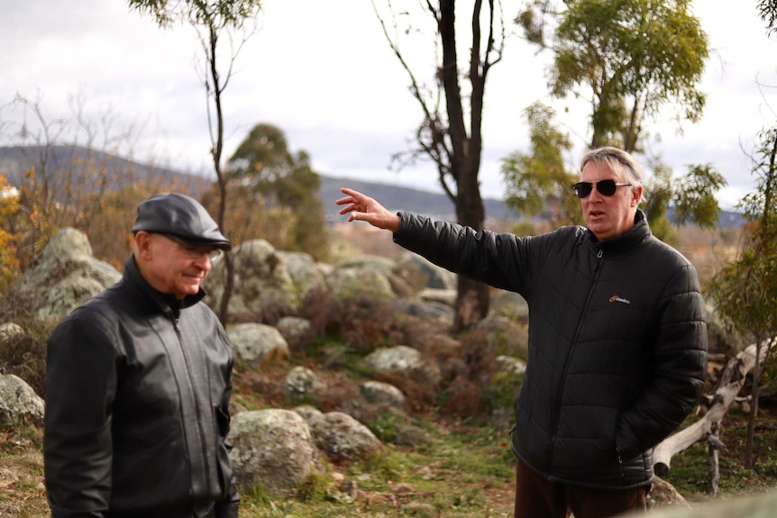 Two men stand amidst a natural landscape chatting.