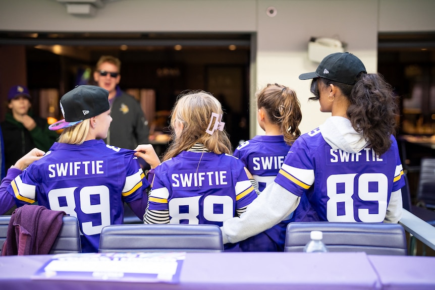 Three girls point to the backs of blue jerseys with Swiftie 89 on them 