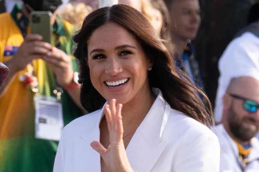 A close up of Meghan Markle smiling and waving at the camera, walking through a crowd of people