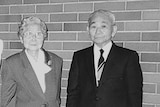 Two elderly people stand in front of a brick wall, in a black and white photograph.