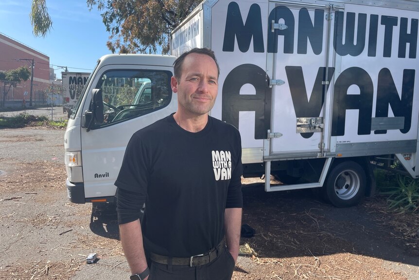 A man wearing all black stands in front of a white removalist van with Man with a Van written on the side