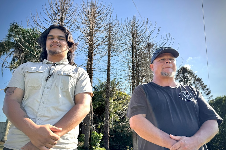 A young indigenous man and his father stand tall with dead Bunya pines behind them