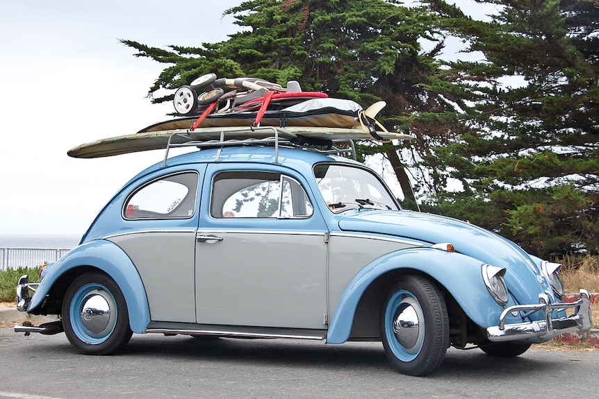 A blue and grey Volkswagen Beetle is pictured with surfboards and pram on its roof.
