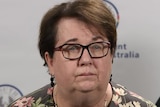 A woman wearing a floral shirt, black glasses and short brown hair stands with a serious expression
