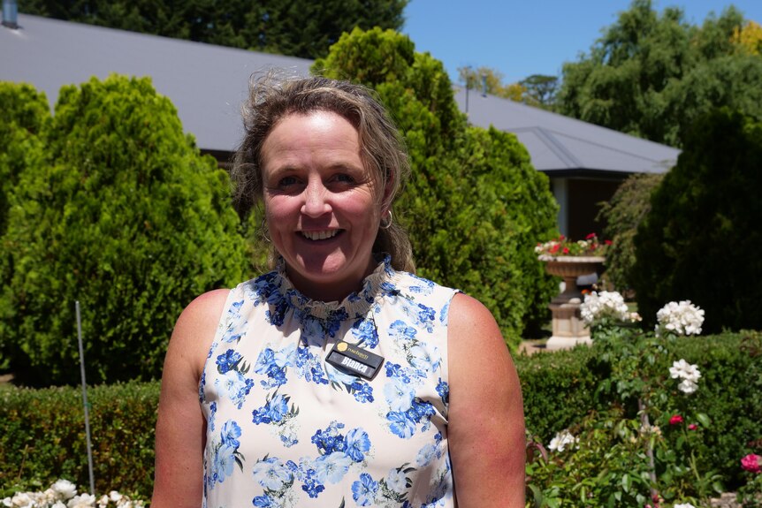 Middle-aged woman wearing blue floral top stands smiling in a rose garden.