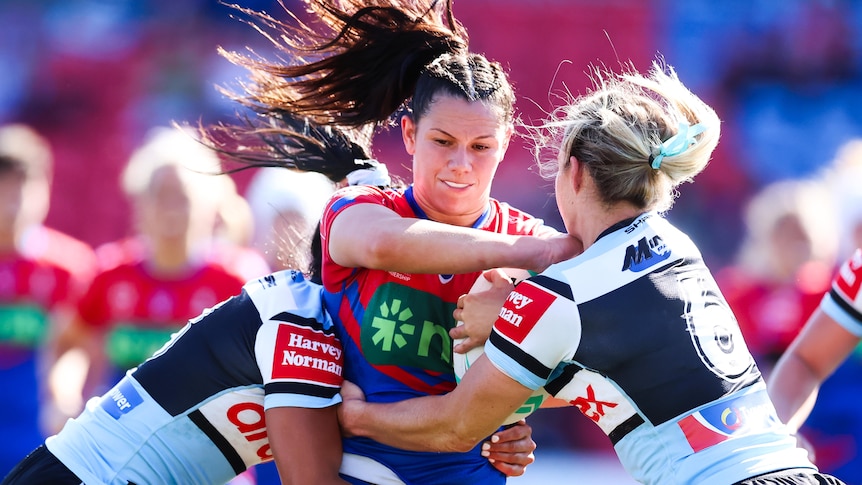 A Newcastle NRLW player grimaces as she holds on to the ball while being tackled by two Cronulla players.