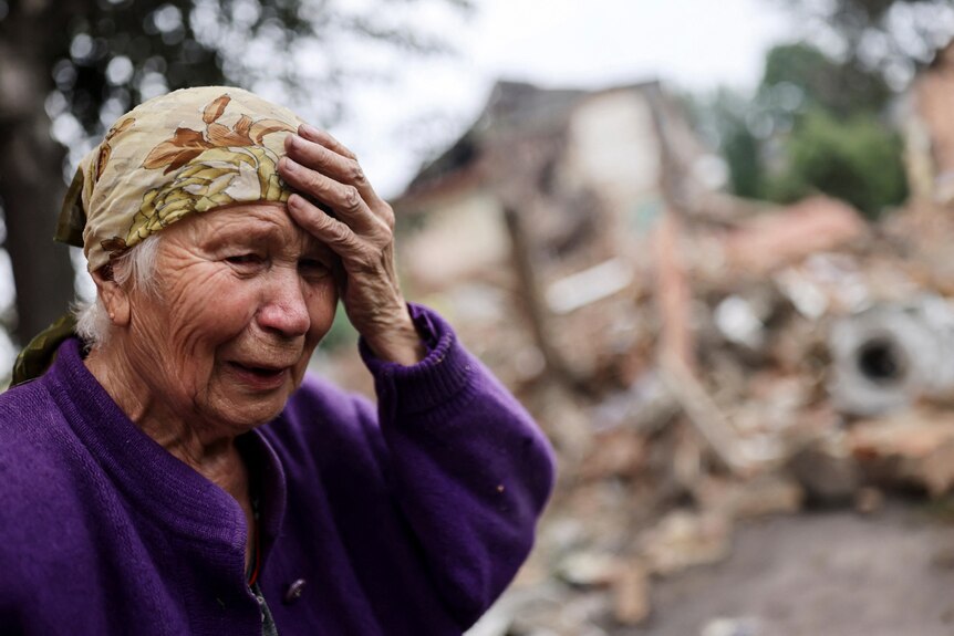 Close up of elderly woman in distress, holding her hand up to her head as she sees damaged buildings.