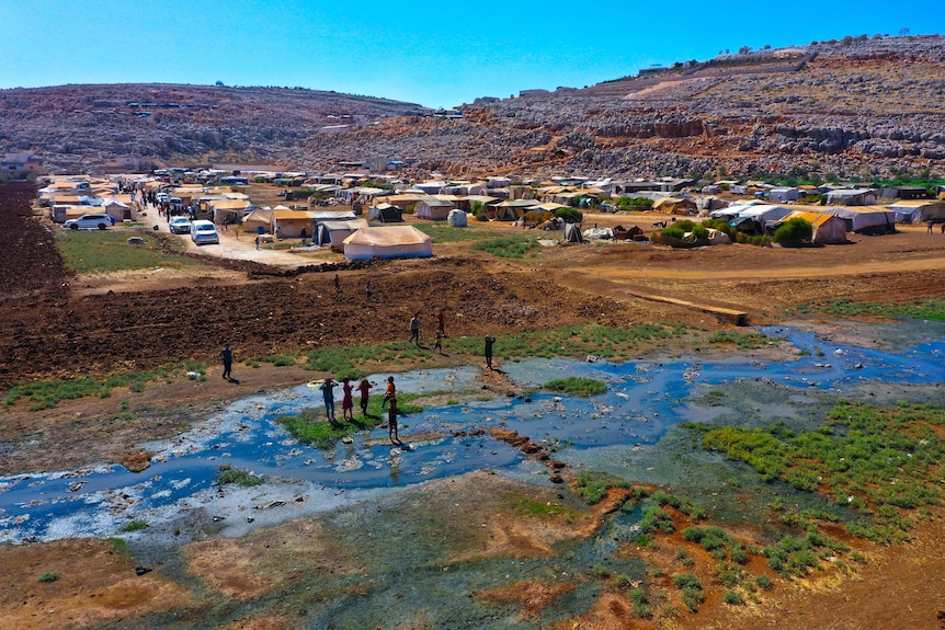 Children play in a shallow stream as a tent city stretches to the horizon behind them. 