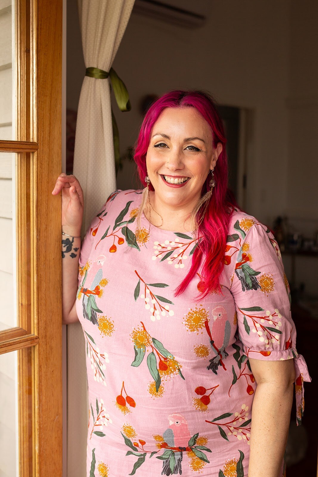 A woman with pink hair and a pink and floral dress stands in a door way smiling. 