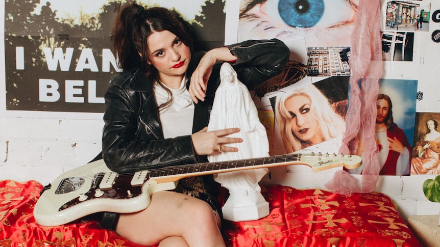 Press shot of Tired Lion's Sophie Hopes; holding guitar, sitting on bed, wearing leather jacket