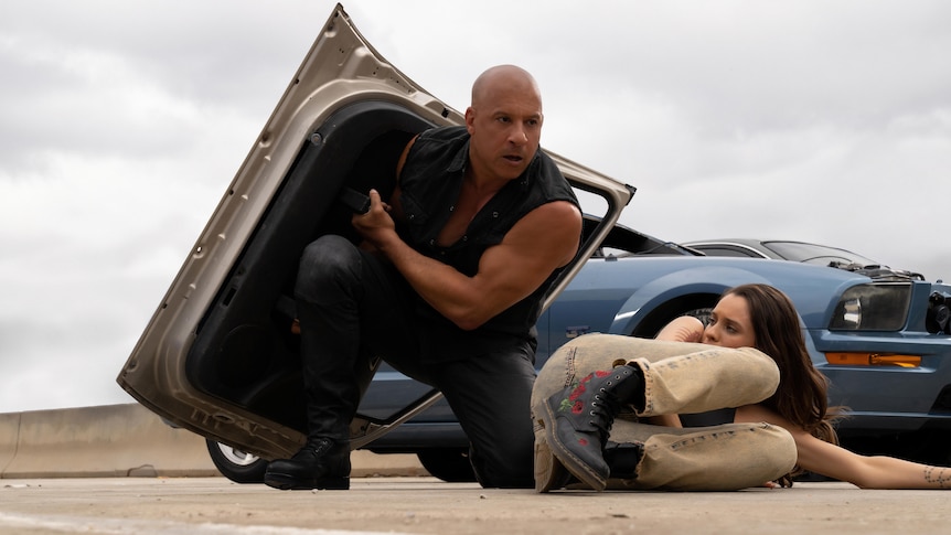 A muscly black man in black clothes holds a car door as a shield over a white woman with brown hair lying on the road.