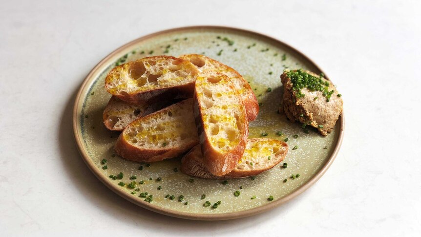A plate of sliced baguette accompanied by a scoop of porcini butter. The snack plate is covered in olive oil and chopped chives.