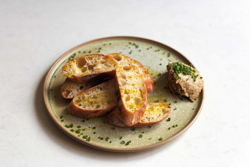 A plate of baguette slices accompanied by a ball of porcini butter.  The snack plate is covered with olive oil and chopped chives.