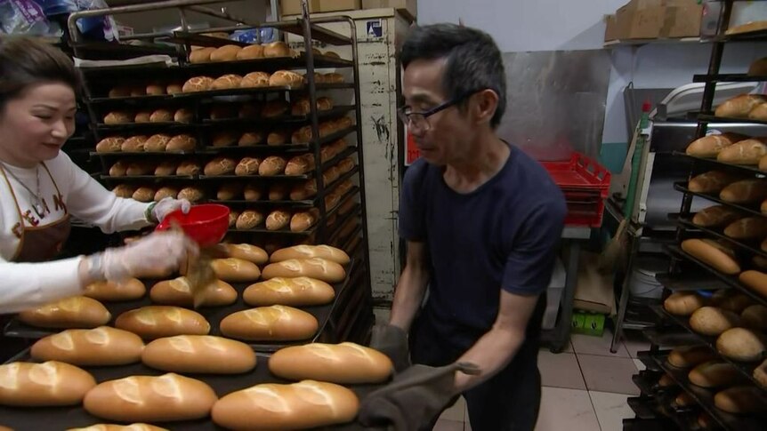A man and a woman tend to bread roles on a baking tray. There are shelves of bread rolls behind them.