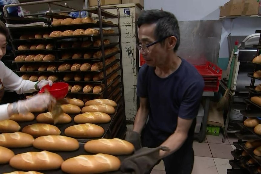 A man and a woman tend to bread roles on a baking tray. There are shelves of bread rolls behind them.
