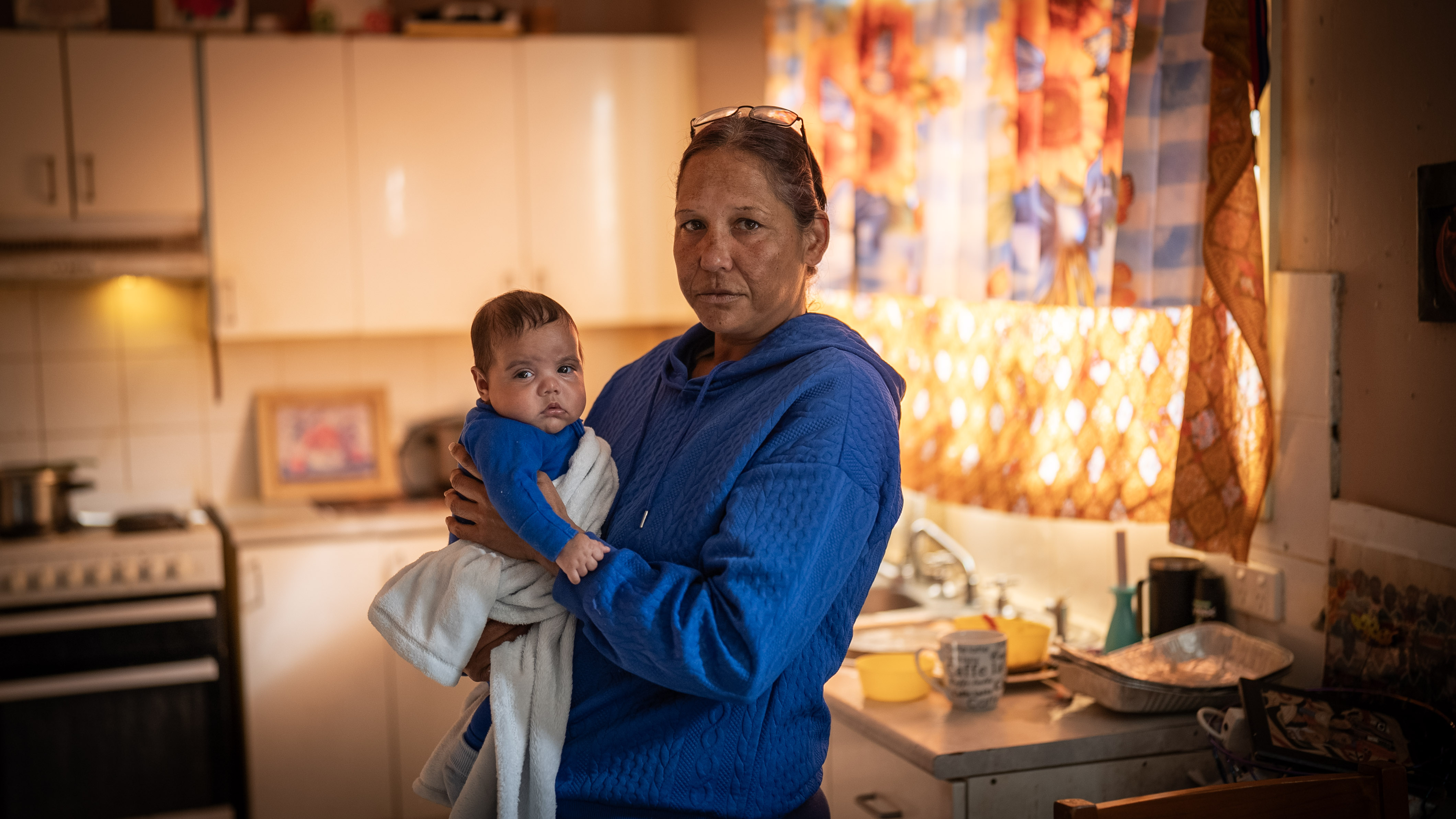 An Indigenous stands in a dimly-lit kitchen holding a baby.