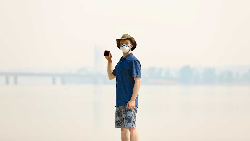 A man stands wearing a mask in thick smoke, holding an air quality monitor.