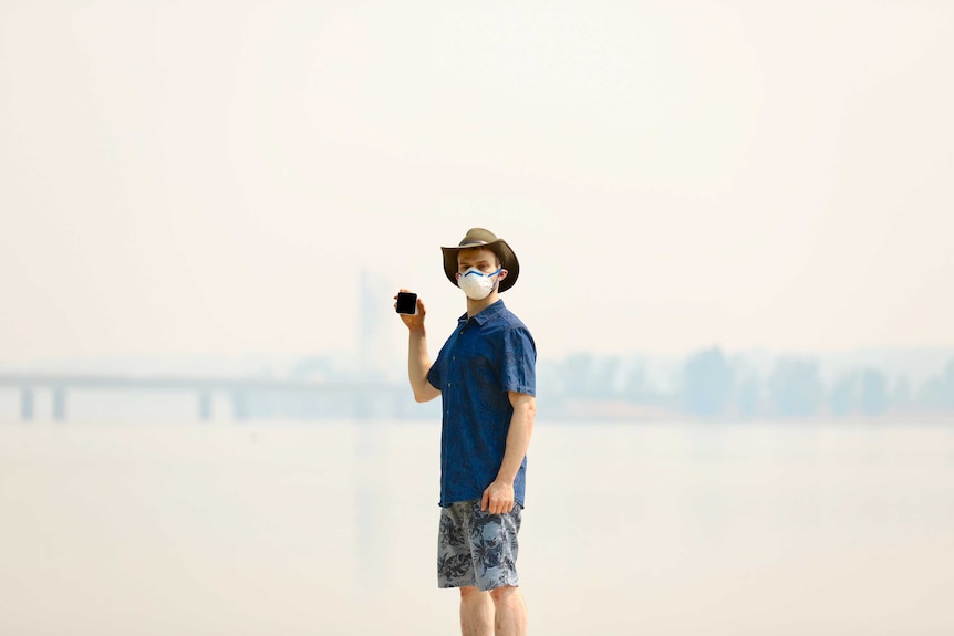 A man stands wearing a mask in thick smoke, holding an air quality monitor.